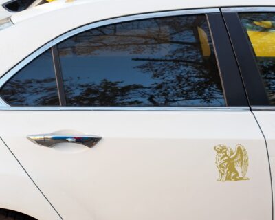 Vinyl decal of a majestic golden retriever dog with large angel wings. The dog is depicted sitting upright with a calm and serene expression, its head slightly tilted upward as if looking toward the heavens. Detailed fur markings create a textured, realistic look throughout the body, while the strong wings extend gracefully from its back, showcasing layered feathers with intricate line work.